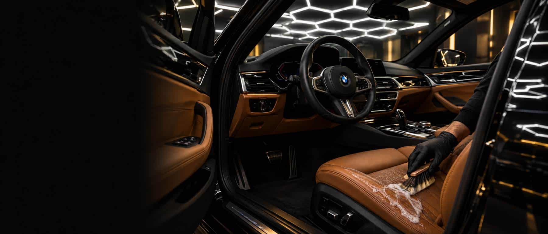 Gehandschoende hand reinigt een cognac lederen zetel van een BMW met een zachte borstel, onder het honingraatlicht